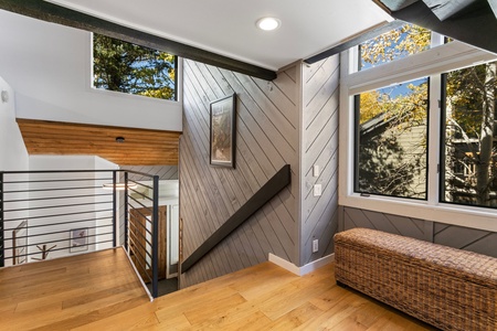 A modern hallway with wooden floors, large windows, a metal railing, a wicker bench, and a framed picture on a wood-paneled wall. A staircase descends to the lower level.