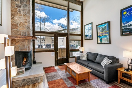 A cozy living room with a stone fireplace, gray sofa, wooden coffee table, and large windows overlooking an outdoor view. Two posters and a lamp can be seen on the walls.