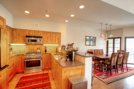 A modern kitchen with wooden cabinets, stainless steel appliances, and a granite countertop. Adjacent is a dining area with a wooden table and chairs, illuminated by hanging lights.