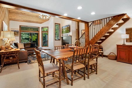 Dining area with wooden table and chairs, adjacent to a living room with a sofa and TV, and a staircase leading to the upper floor. Large windows offer a view of greenery outside.