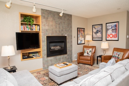 A living room with a wall-mounted TV, a fireplace, two armchairs, a sofa, and an ottoman. It features framed posters on the walls, a patterned rug, and a bookshelf with plants and books.