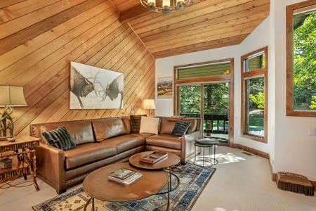 A cozy living room with a high slanted wood-paneled ceiling, a brown leather sectional sofa, a large nature-themed artwork, and large windows offering a view of green trees.