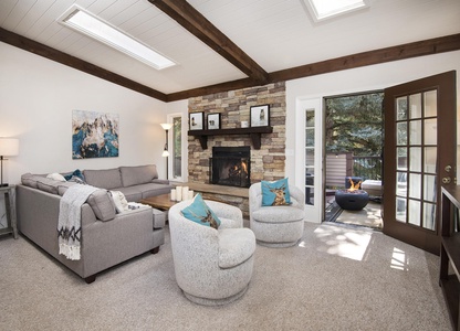 A cozy living room with a stone fireplace, gray sectional sofa, two gray swivel chairs, and a coffee table. There's an open door leading to an outdoor area with seating and a fire pit.