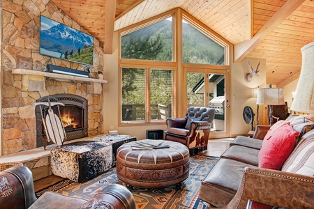 A cozy living room with a stone fireplace, wooden ceiling, large windows, a leather armchair, an ottoman, patterned rugs, and a TV mounted above the fireplace. Sunlight streams through the windows.