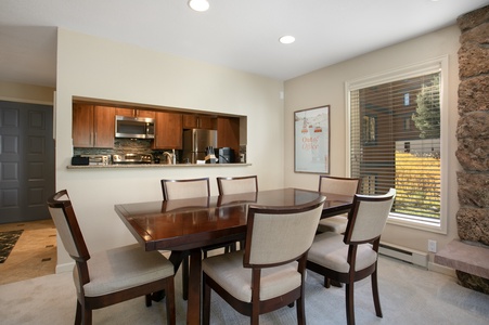 A dining room with a wooden table and six chairs, adjacent to a kitchen with wooden cabinets and stainless steel appliances, and a window showing an outdoor view.