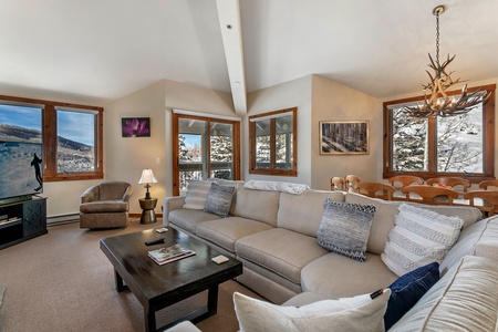 A spacious living room with large windows, beige sectional sofa, dark wooden coffee table, a TV, and mountain view. The room is decorated with cushions, antler chandelier, and wall art.