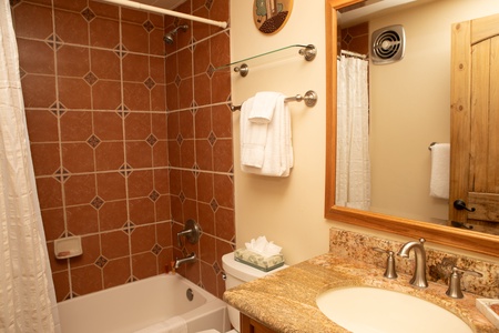 Bathroom with a tiled bathtub and shower combo, beige countertop sink, wooden door, mirror, towel racks with white towels, and a tissue box on the toilet tank.