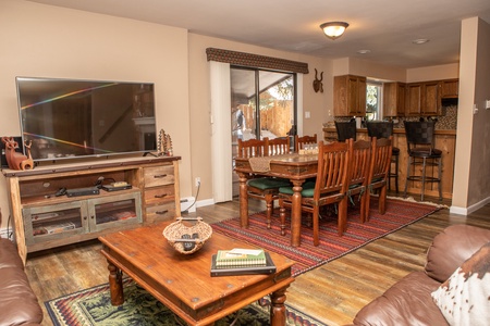 A cozy living and dining area featuring a flat-screen TV on a wooden stand, a dining table with eight chairs, and a kitchen counter with bar stools. The room has wooden floors and a rug.