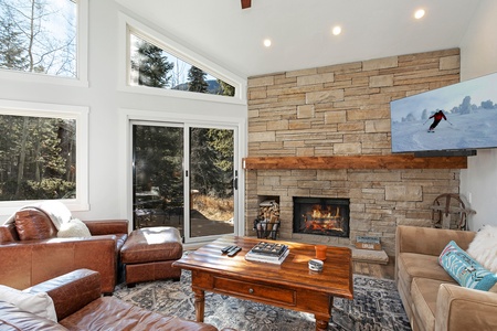 A cozy living room featuring a stone fireplace, a mounted TV displaying a skier, large windows with treetop views, and leather seating around a wooden coffee table.