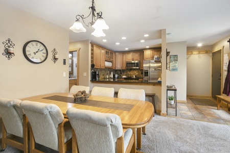 Dining and kitchen area in a house with a wooden table and six upholstered chairs, a wall clock, ceiling lights, and a modern kitchen with stainless steel appliances and wooden cabinets.