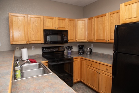 A modern kitchen with wooden cabinets, a black microwave, a black electric stove, a black refrigerator, a double sink with a soap dispenser, and a countertop with kitchen utensils and appliances.