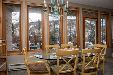 A dining area with a glass table, six yellow chairs, and a blue chandelier. Large windows offer a snowy mountain view.