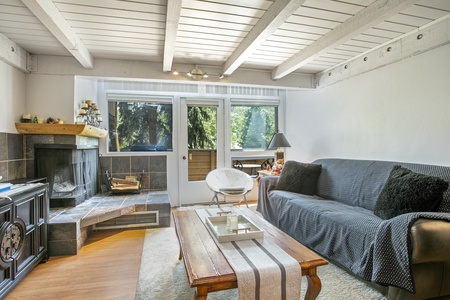 A living room with exposed wooden beams, a grey sofa, a wooden coffee table, a fireplace, and large windows providing a view of greenery outside.