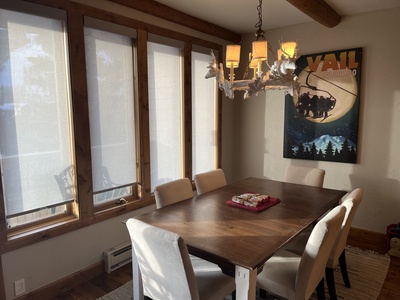 A wooden dining table with six white chairs in a room with large windows and a vintage "Vail Colorado" poster on the wall. A chandelier with a rustic design hangs above the table.