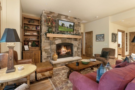 A cozy living room with a stone fireplace, a flat-screen TV above it, and various furniture items including a maroon sofa, armchairs, a wooden coffee table, and a round side table with a lamp.