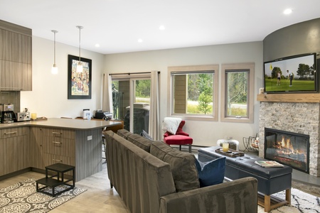 A modern living room with a gray couch, a TV above a fireplace, a kitchen area with wooden cabinets, and a sliding door leading to an outdoor view.