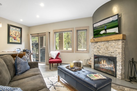 A cozy living room with a stone fireplace, TV mounted above it, sofa, red chair, and windows. A colorful poster and a sliding door leading to a balcony are on the left wall.