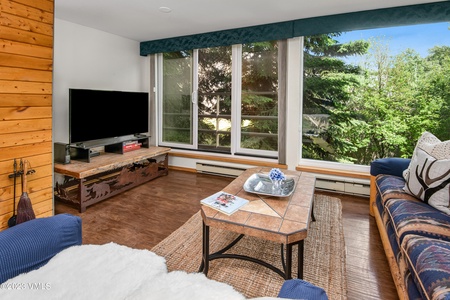 A living room with large windows overlooking trees, featuring a television on a wooden stand, a tile-topped coffee table, a patterned sofa, and wooden flooring.