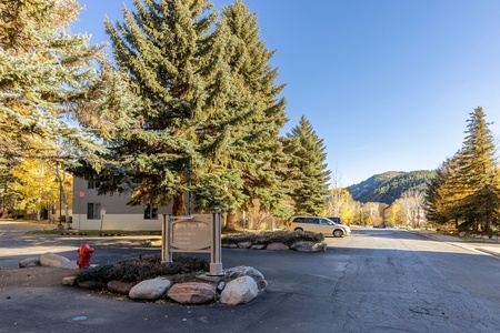 A parking lot with a sign and a red fire hydrant under tall evergreen trees. In the background, a building, parked cars, and tree-covered hills under a clear blue sky.