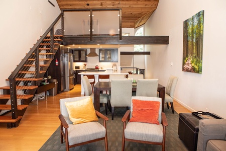 A modern living room and kitchen area with lofted bedroom. Contains chairs, dining table, kitchen appliances, stairs to loft, and forest-themed wall art.