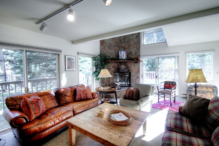 A cozy living room with large windows, brown leather sofa, a wooden coffee table, armchairs, and a stone fireplace. Sunlight streams in, and there is a view of trees outside.