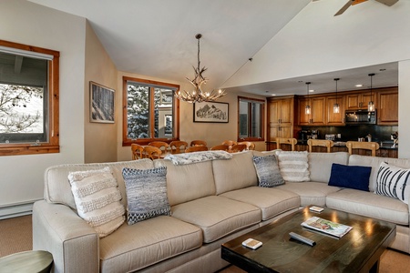 A spacious living room features a large beige sectional sofa with patterned cushions, a wooden coffee table, and a view of the open dining area and kitchen with wooden cabinetry in the background.