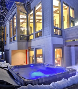 A modern house with large windows is lit from within. Outside, there is a hot tub with blue lighting, surrounded by snow.