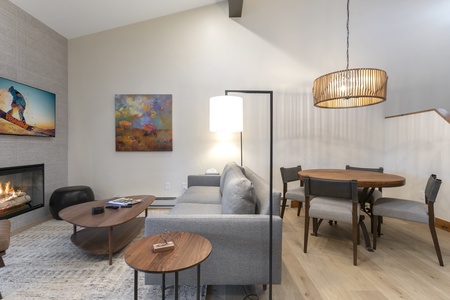 A modern living room and dining area with a gray sofa, round wooden table with four chairs, wall-mounted TV, colorful artwork, and a lit fireplace. A pendant light hangs above the dining table.