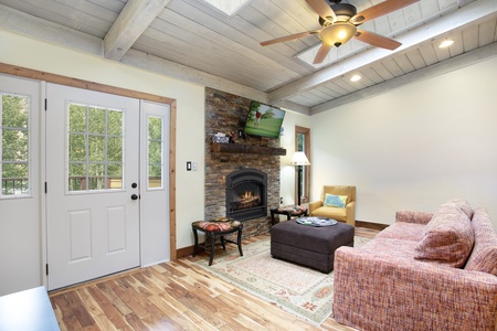 A living room with a ceiling fan, stone fireplace, wall-mounted TV, sofa, and ottoman. Large windows and door on the left side.