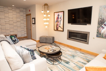 A modern living room with a white sectional sofa, two grey ottomans, a round coffee table, wall art, a mounted TV above a fireplace, and decorative lighting.