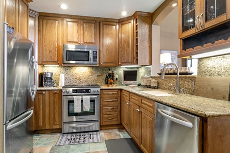 Contemporary kitchen with stainless steel appliances, wooden cabinets, granite countertops, and a tiled floor. A tea towel hangs on the oven door, and a rug near the sink says "home sweet home.