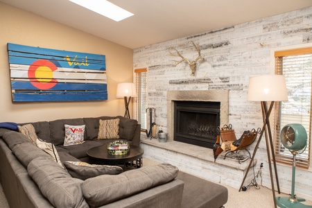 A cozy living room features a sectional sofa, round coffee table, and a fireplace with a wood-paneled accent wall. The walls display art, including a piece with the Colorado flag. Two floor lamps provide light.