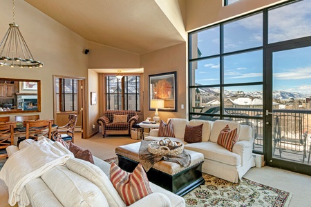 A spacious living room features large windows with a mountain view, a white sectional sofa, an ottoman with decorations, a rug, and a dining area with a kitchen visible in the background.