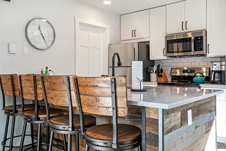 Modern kitchen with a large island featuring a marble countertop, wooden bar stools, stainless steel appliances, a digital clock on the wall, and a roll of paper towels on the counter.