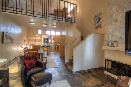 A cozy living area with a fireplace, black leather chair, hardwood flooring, an adjacent kitchen with wooden cabinetry, and an upstairs loft. A dining table and chairs are set near the kitchen.