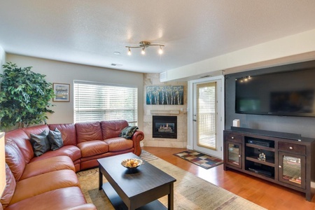 A living room with a red sectional sofa, a coffee table, a wall-mounted TV, a fireplace, a large plant, and windows with blinds. There is also a door leading outside and a rug on the hardwood floor.