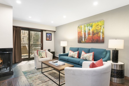 A modern living room with a blue sofa, two white chairs, a coffee table, a fireplace, and a large sliding door to a balcony. There is a colorful painting above the sofa and two table lamps.