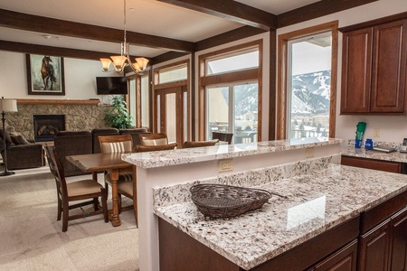 A modern kitchen with a granite countertop, a wicker basket, and dark wood cabinetry. Adjacent dining area with a wooden table and leather chairs, and a living room with a stone fireplace and mountain view.