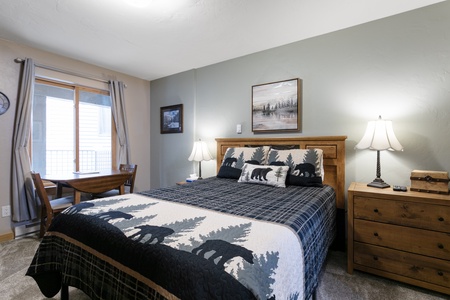 A neatly arranged bedroom with a wooden bed framed with bear-themed bedding, two bedside lamps, a wooden nightstand, a small table and chairs by the window, and two landscape pictures on the walls.