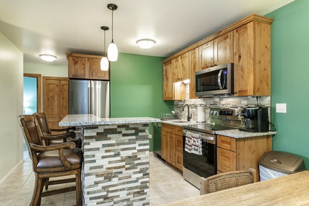 A modern kitchen with wood cabinets, granite countertops, stainless steel appliances, an island with stone accents, and bar stools. The walls are green and the floor is tiled.