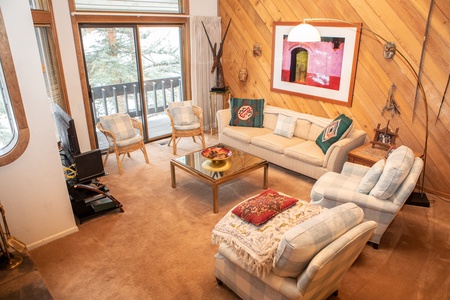 A cozy living room features beige sofas, a glass coffee table, and plaid armchairs. Art decorates a wood-paneled wall, while a sliding glass door leads to a snow-covered balcony outside.