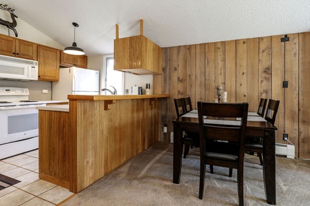 A kitchen and dining area featuring wooden cabinets, a stove, and a microwave. The dining space includes a dark wooden table with four chairs and is adjacent to wooden wall paneling.