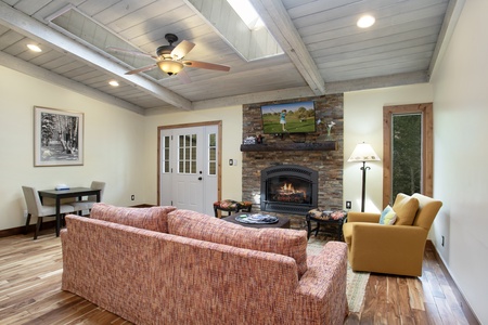 Living room with a vaulted ceiling, ceiling fan, fireplace, and a mix of modern and rustic decor. A sofa and armchair face the fireplace, and a small desk is in the corner.