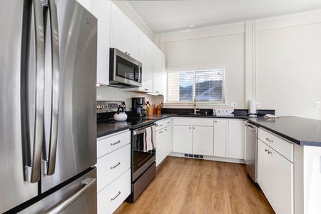 Modern kitchen with stainless steel appliances, white cabinetry, a black countertop, and wooden flooring. A window above the sink provides natural light.