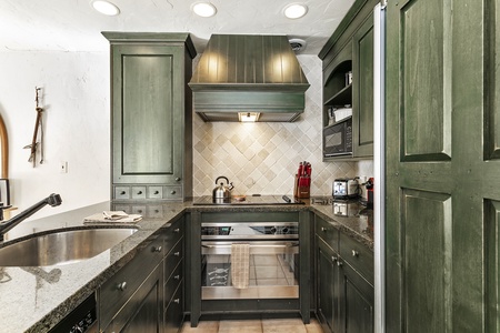 A compact kitchen featuring green cabinetry, granite countertops, a stainless steel oven, a stovetop with a range hood, a sink, and various appliances including a kettle and a knife set.