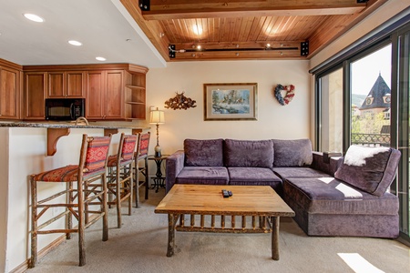 A cozy living room with a purple sectional sofa, wooden coffee table, and a kitchen bar with four cushioned stools. The ceiling has wooden beams and a large window provides natural light.