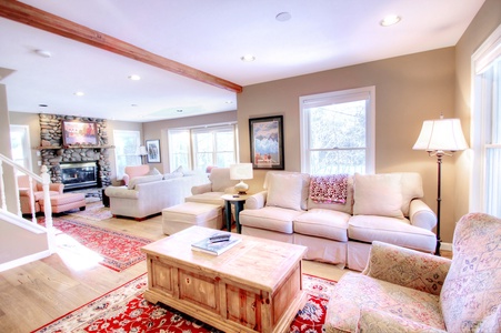 A brightly lit living room with beige sofas, a wooden coffee table, and a stone fireplace. A red rug adds color, and lamps provide additional light. Multiple windows allow natural light to flood the room.