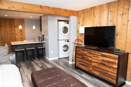 A cozy room with wooden walls, a TV on a dresser, a washer and dryer stack, and a kitchen area with bar stools and pendant lights.