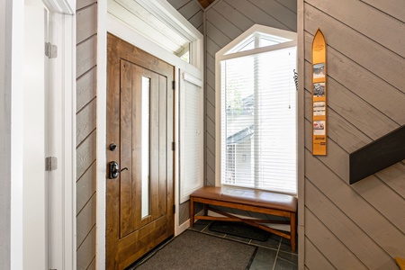 A wooden front door with glass panels, a side window, a bench beneath another window, and a narrow wall-mounted shelf with decorations in a home entryway.