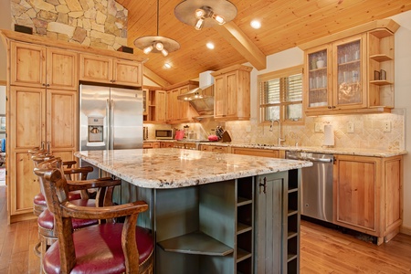 A spacious kitchen with wooden cabinets, a stainless steel refrigerator, and a large island with a marble countertop. Three wooden bar stools with red cushions are placed at the island.